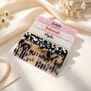 May include: A stack of five rectangular hair combs made of marbled acrylic in various colors including pink, white, and black. Each comb features a personalized name or title printed in script font, specifically Isabella, bridesmaid, Taylor, Bride, and Hailey.