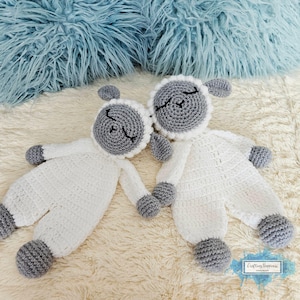 May include: Two handmade crochet lamb dolls. The lambs are white with gray faces, ears, and feet. They are lying on a cream-colored surface, with a blue fluffy background. The lambs are holding hands.