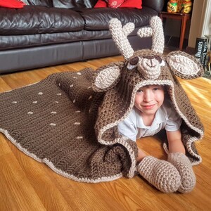 May include: A child wearing a brown and beige crocheted deer hooded blanket with a white shirt. The blanket has a deer face with a brown nose and white spots.