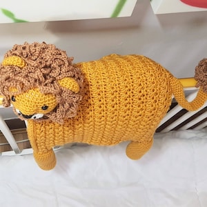 May include: A crocheted lion toy with a brown mane, yellow body, and a brown tail. The toy is hanging from a white crib rail.