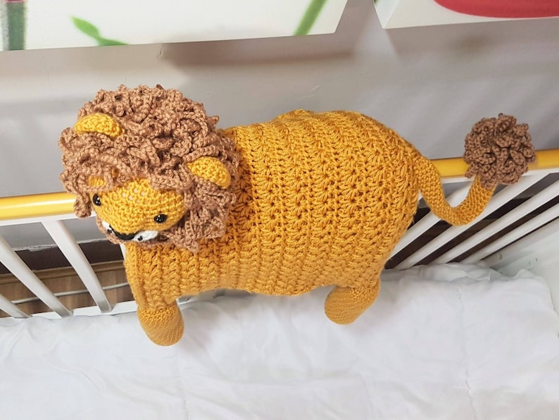 May include: A crocheted lion toy with a brown mane, yellow body, and a brown tail. The toy is hanging from a white crib rail.