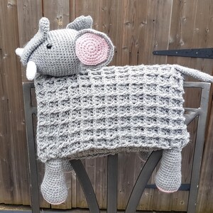 May include: A gray crocheted elephant blanket with pink ears and a white trunk. The blanket is draped over a black metal gate.