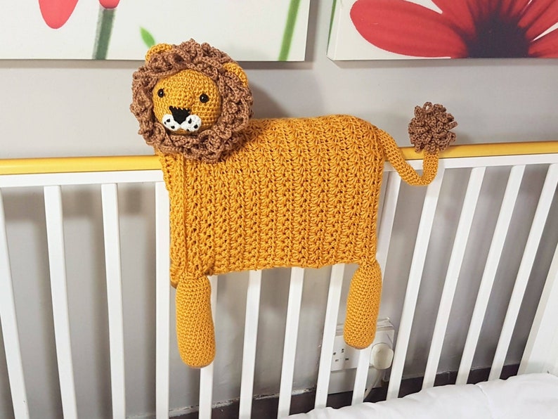 May include: A crocheted lion baby blanket with a brown mane and tail. The blanket is yellow and is hanging over a white crib.