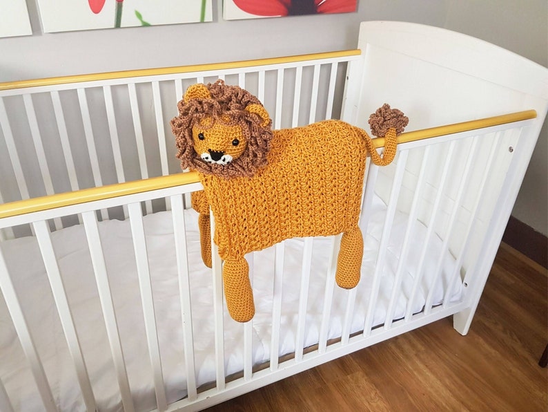 May include: A crocheted lion toy with a brown mane and a yellow body. The toy is hanging over the side of a white crib.