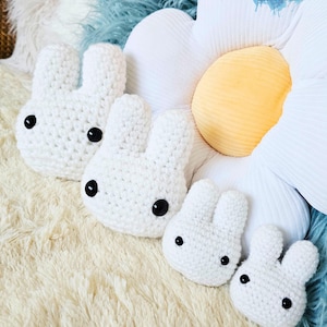May include: Four white crochet bunny heads with black button eyes are arranged on a fluffy, cream-colored surface. The bunnies vary in size, suggesting a family or set. A large daisy-shaped pillow with a yellow center and white petals is in the background.