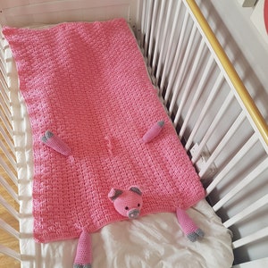 May include: A pink crocheted blanket with a pig design. The blanket features a pig face in the centre and two pig legs on each side. The blanket is lying on a white cot mattress. Pattern by Crafting Happiness