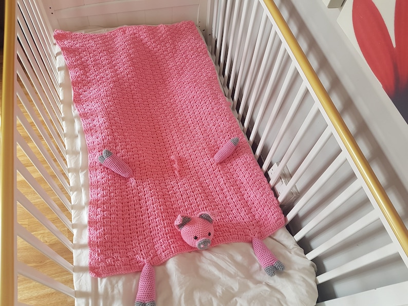 May include: A pink crocheted blanket with a pig design. The blanket features a pig face in the centre and two pig legs on each side. The blanket is lying on a white cot mattress. Pattern by Crafting Happiness