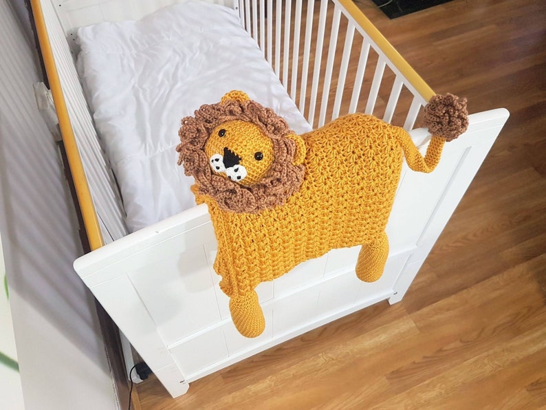 May include: A crocheted lion toy in yellow and brown with black eyes and a white nose. The toy is hanging over the side of a white crib.