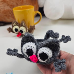May include: A dark gray crocheted cat mug hug with large white and black eyes and a bright pink nose. The cat has four small limbs extending outward. In the background, a brown crocheted owl mug hug is wrapped around a yellow ceramic mug.