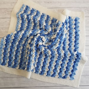 May include: A blue and white crocheted baby blanket with a textured, raised pattern. The blanket is folded and laid on a light wood surface.