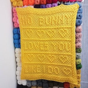 Puede incluir: Una manta de crochet amarilla con un patrón texturizado. La manta está colgada en una pared con ovillos de lana de varios colores detrás.