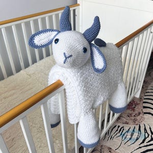 Puede incluir: Una cabra de ganchillo blanca hecha a mano con detalles azules, incluyendo cuernos, orejas y pezuñas. El peluche tiene ojos azules de botón y una boca cosida. La cabra está colocada sobre la barandilla blanca de una cuna.