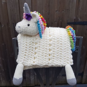 Könnte beinhalten: Eine weiße gehäkelte Einhorn-Decke mit einer Regenbogenmähne und einem Regenbogen-Schwanz. Das Einhorn hat ein graues Horn und eine lila Blume auf dem Kopf.