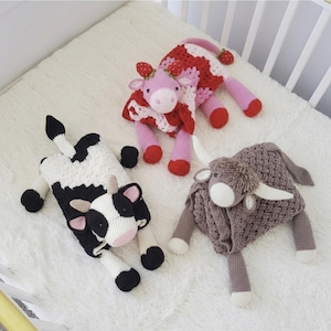 Può includere: Tre peluche a forma di mucca fatti a mano all'uncinetto. Uno è bianco e nero, un altro rosso e bianco con dettagli di fragole, e il terzo è una mucca delle Highlands marrone. I giocattoli sono su una superficie bianca e soffice.