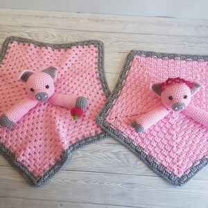 May include: Two pink crocheted baby blankets with grey borders. Each blanket features a crocheted pig toy with a pink nose and black eyes. One pig has a red flower on its head. Pattern by Crafting Happiness