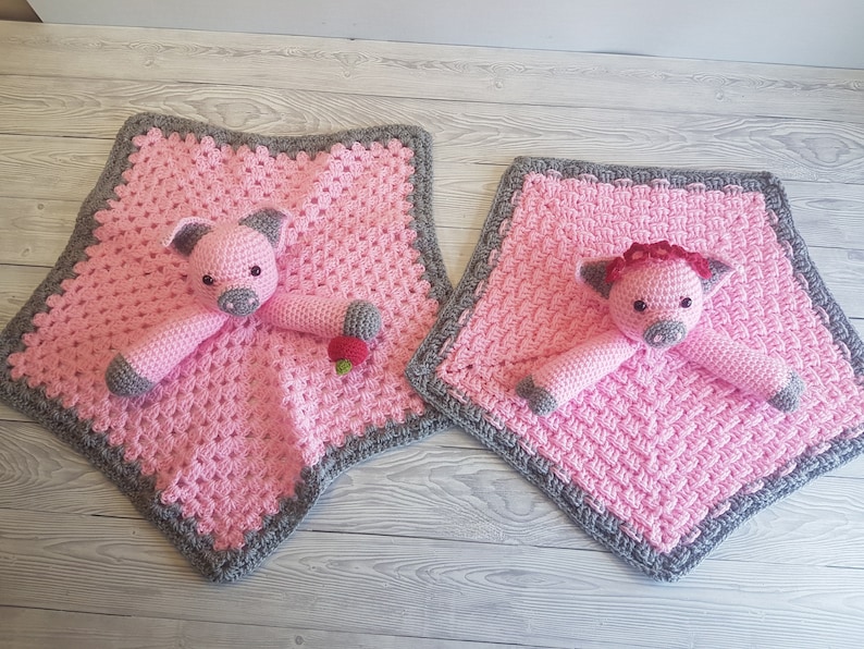 May include: Two pink crocheted baby blankets with grey borders. Each blanket features a crocheted pig toy with a pink nose and black eyes. One pig has a red flower on its head. Pattern by Crafting Happiness