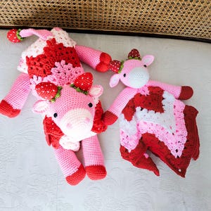 May include: Two crocheted stuffed cows, one larger than the other, are both pink with red and white accents. The larger cow has a strawberry pattern on its body. The smaller cow is holding a blanket with a strawberry pattern.