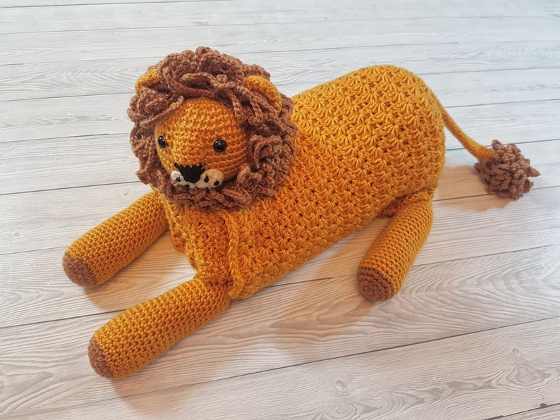 May include: A crocheted lion stuffed animal with brown mane and a golden body. The lion is lying on a light wood surface.