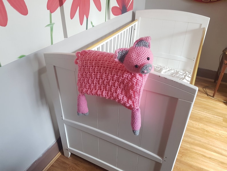 K&ouml;nnte beinhalten: Ein rosa geh&auml;keltes Schweinchenspielzeug mit grauen Ohren und Schnauze, das an einem wei&szlig;en Babybett h&auml;ngt.