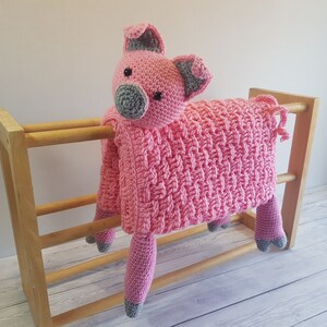 May include: A pink crocheted pig blanket with grey accents. The blanket is draped over a wooden rack. Pattern by Crafting Happiness