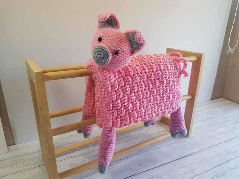 May include: A pink crocheted pig blanket with grey accents. The blanket is draped over a wooden rack. Pattern by Crafting Happiness