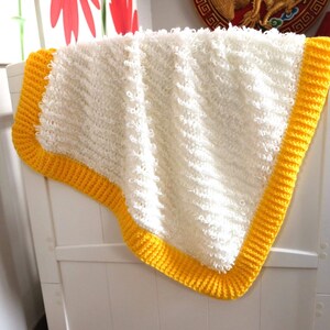Puede incluir: Manta de bebé de crochet blanco con un borde de crochet amarillo. La manta está doblada y drapeada sobre una cuna blanca.