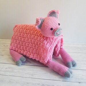 May include: A pink crocheted pig toy with grey accents. The pig is lying on its side with its legs outstretched. Pattern by Crafting Happiness