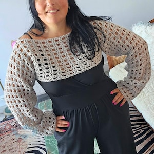 Op de afbeelding: Een beige gehaakte bolero met lange mouwen. Het open ontwerp van de bolero is zichtbaar. Het kledingstuk wordt gedragen over een zwarte top en een zwarte broek. De mouwen hebben een lichte klokvorm bij de pols.