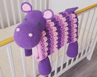 May include: A purple and pink crocheted hippopotamus toy. The hippopotamus is sitting on a white crib.