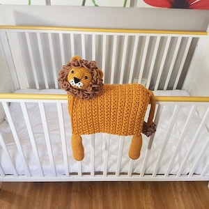 May include: A crocheted lion toy with a brown mane and a white face, hanging over the side of a white crib.