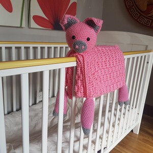 May include: A pink crocheted pig blanket with grey accents hanging over a white cot. The blanket is soft and cuddly, perfect for a baby's nursery. Pattern by Crafting Happiness