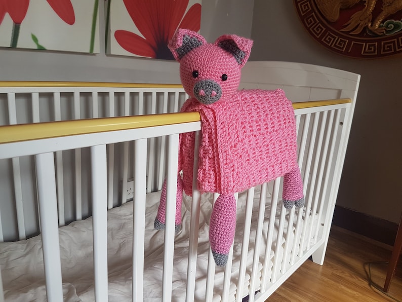 May include: A pink crocheted pig blanket with grey accents hanging over a white cot. The blanket is soft and cuddly, perfect for a baby's nursery. Pattern by Crafting Happiness