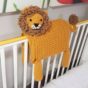 May include: A crocheted lion toy with a brown mane and yellow body. The toy is hanging over a white crib with a wooden rail.