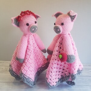 May include: Two pink crocheted pig stuffed animals with grey accents. One pig has a red flower on its head and the other pig is holding a pink flower. Pattern by Crafting Happiness