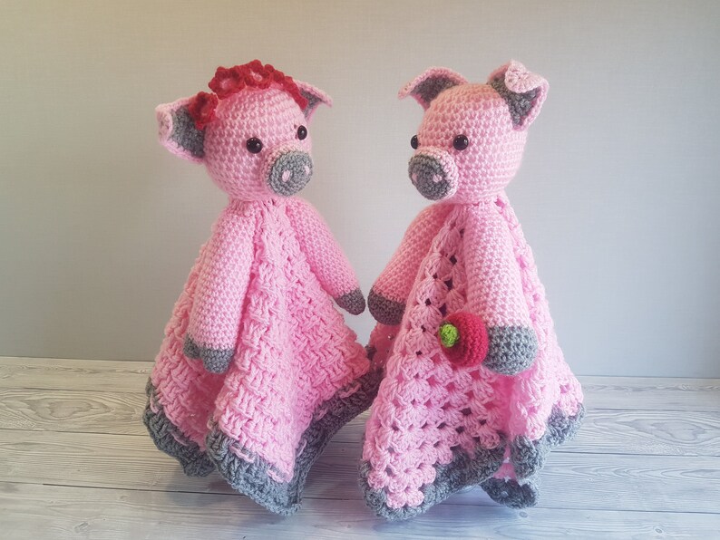 May include: Two pink crocheted pig stuffed animals with grey accents. One pig has a red flower on its head and the other pig is holding a pink flower. Pattern by Crafting Happiness