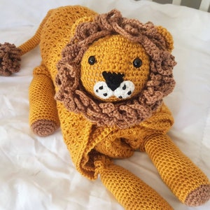 May include: A crocheted lion stuffed animal with a brown mane and yellow body. The lion is lying on a white surface.