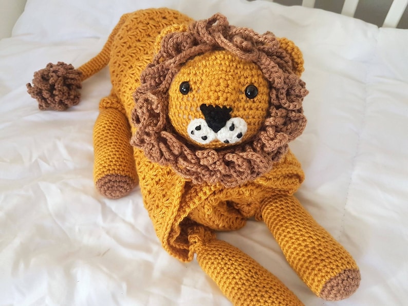 May include: A crocheted lion stuffed animal with a brown mane and yellow body. The lion is lying on a white surface.