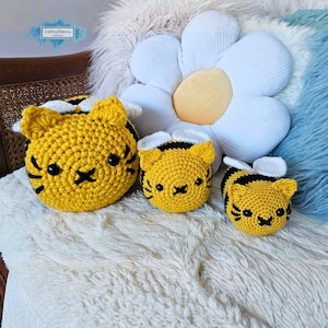 May include: Three handmade crochet bee-themed items in varying sizes. The bees are yellow with black stripes and facial details, including black eyes and a stitched mouth. White wings are attached to the top of each bee. The items are displayed on a white, fluffy surface.