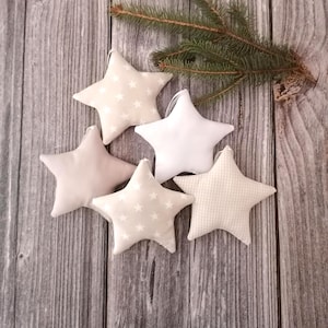 Könnte beinhalten: Fünf Stoffstern-Ornamente mit Aufhängeschlaufen. Zwei Sterne haben einen beigen Hintergrund mit weißen Sternen, einer ist einfarbig weiß, einer einfarbig beige und einer hat ein beigeweißes Vichy-Karo-Muster. Jeder Stern ist weich gepolstert.