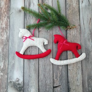 May include: Two handmade rocking horse ornaments. One is white with a red base and a pink bow, the other is red with a white and red base and a pink bow. The ornaments are hanging from a green pine branch against a wooden background.