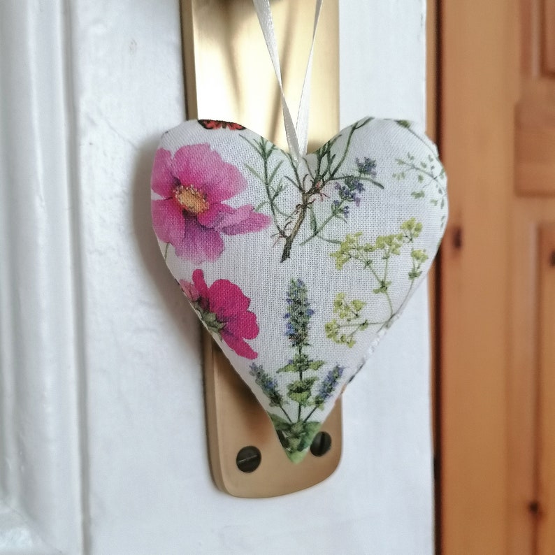 May include: A heart-shaped fabric ornament featuring a botanical print with pink flowers, green herbs, and small blue blossoms. The heart is suspended by a thin white ribbon from a brass-colored metal door handle plate against a white door frame.