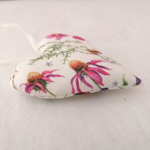 May include: A heart-shaped fabric ornament featuring a cream-colored background with a botanical print of vibrant pink coneflowers, green foliage, and small blue flowers. A thin white ribbon is attached to the top for hanging purposes.