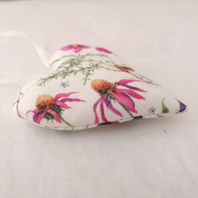 May include: A heart-shaped fabric ornament featuring a cream-colored background with a botanical print of vibrant pink coneflowers, green foliage, and small blue flowers. A thin white ribbon is attached to the top for hanging purposes.