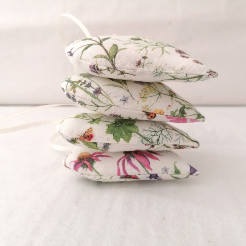 May include: A stack of four heart-shaped fabric ornaments featuring a botanical print with green leaves, yellow, pink, and purple wildflowers, and small butterflies on a cream background, each with a thin white ribbon loop attached for hanging.
