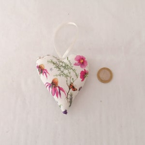 May include: A heart-shaped fabric ornament featuring a white background with a botanical print of pink flowers, green foliage, and a small butterfly. A thin white ribbon loop is attached to the top. A one-euro coin is placed next to it for scale.