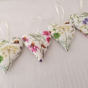May include: Four heart-shaped fabric ornaments featuring a white background with botanical illustrations of yellow and pink flowers, green foliage, butterflies, and snails. Each stuffed heart includes a thin white ribbon loop for hanging purposes.