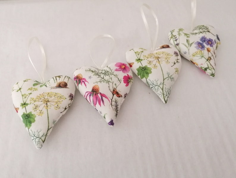 May include: Four heart-shaped fabric ornaments featuring a white background with botanical illustrations of yellow and pink flowers, green foliage, butterflies, and snails. Each stuffed heart includes a thin white ribbon loop for hanging purposes.