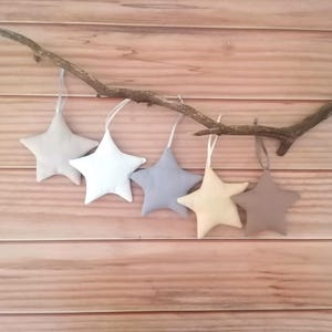 May include: Five plush star-shaped ornaments in varying shades of light gray, white, blue-gray, cream, and taupe. Each star features a matching fabric ribbon loop for hanging and is suspended from a natural wooden branch with a textured bark surface.
