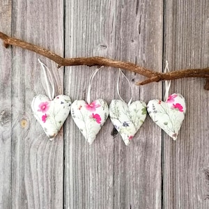 May include: Four stuffed fabric hearts hanging from a natural wooden branch. Each heart features a white background with a botanical print of green stems, leaves, and vibrant pink flowers. Each heart is suspended by a thin white ribbon loop.