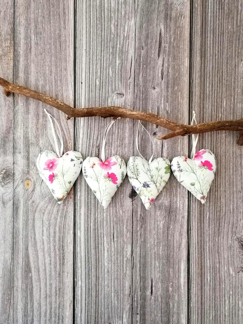 May include: Four stuffed fabric hearts hanging from a natural wooden branch. Each heart features a white background with a botanical print of green stems, leaves, and vibrant pink flowers. Each heart is suspended by a thin white ribbon loop.